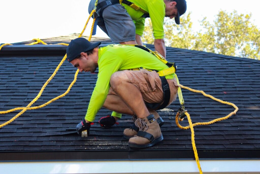 A worker inspects a residential shingle roof under bright sunlight with trees in the background.