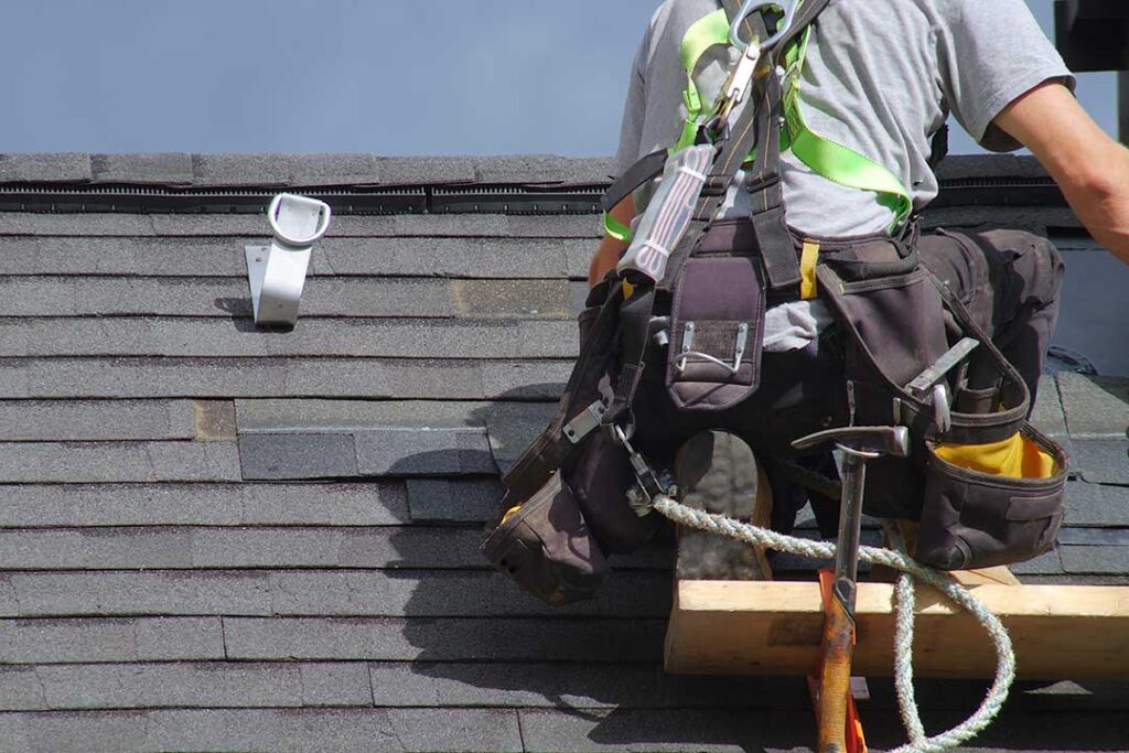 A person inspecting a shingle roof before a storm, with tree branches and gutters in view.